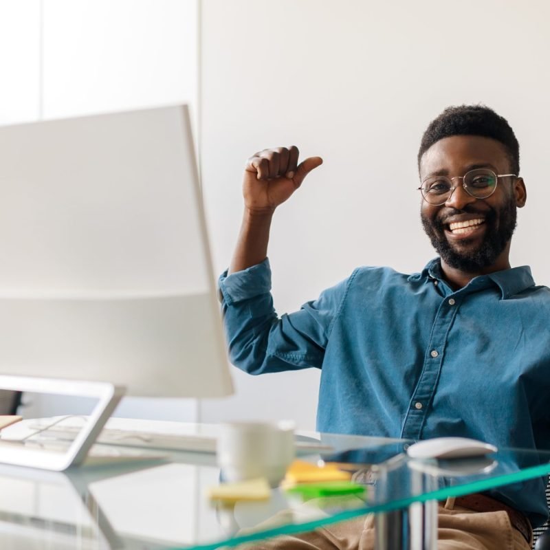Excited black businessman gesturing yes in front of computer, celebrating success, sitting at workplace in office, panorama. Male entrepreneur making great deal, signing online contract